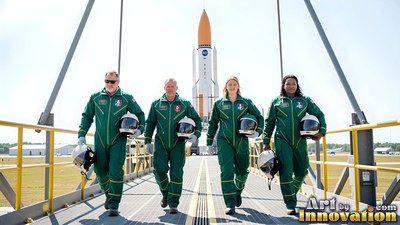 Walking on a walkway, four astronauts in green suits hold helmets before a rocket