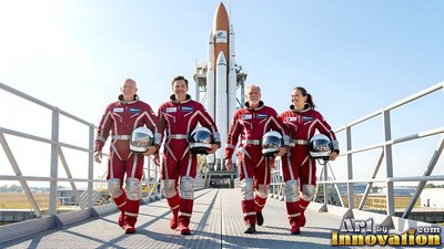 Walking side by side on a walkway, four astronauts wear red flight suits near a rocket