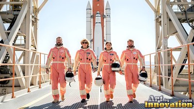 Four astronauts in orange flight suits walk down a sunny launch bridge toward a rocket