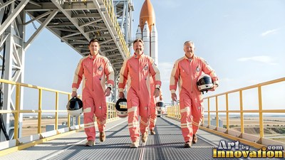 Three astronauts in pink flight suits walk on a sunny launch walkway