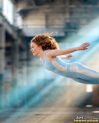 In an empty warehouse, a woman in a light blue jumpsuit leaps horizontally through light