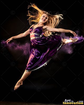 Leaping in the air, a smiling woman dances in a purple dress with purple powder around her
