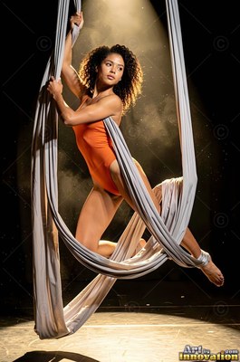 A woman in an orange leotard poses in hanging gray silks under a warm spotlight
