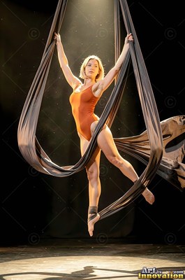 Under a single spotlight, a woman in an orange leotard hangs from silk ribbons