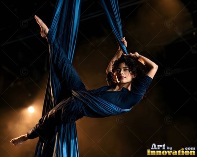 A woman in a dark blue top hangs from blue aerial silks against a dark, warm background