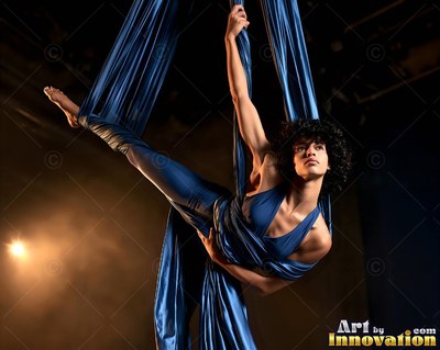 A woman hangs suspended in blue aerial silks with warm spotlight behind her