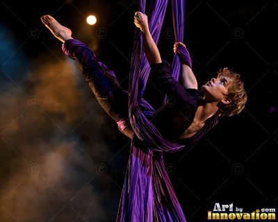 A woman in dark clothes hangs midair wrapped in long purple silks