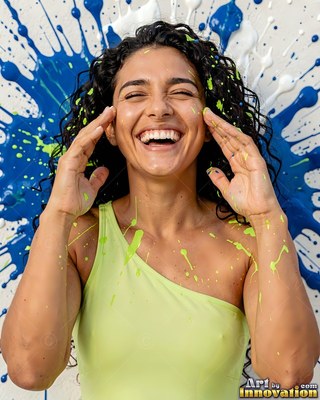 In front of blue paint splatters, a grinning woman in a yellow top laughs