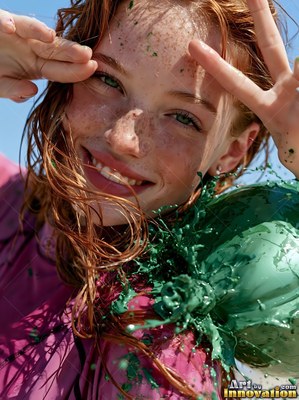 Freckles cover the smiling red-haired woman as thick green paint splatters over her shoulder outdoors