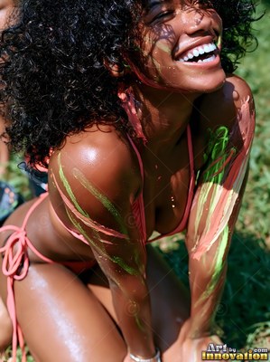 Kneeling on green grass, a laughing woman wears a pink bikini smeared with paint