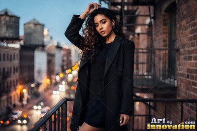 A woman holding the rail of a fire escape among the wooden water towers, the avenue lights coming on far below at blue hour