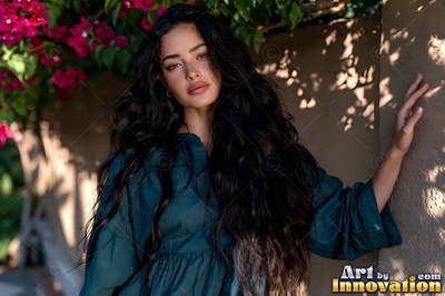 Long dark wavy hair falls past a woman in teal leaning against a sunny wall under pink flowers