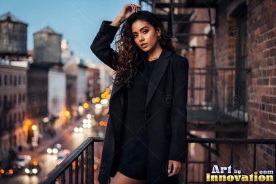 A woman in a black coat stands on a balcony above a busy street at dusk