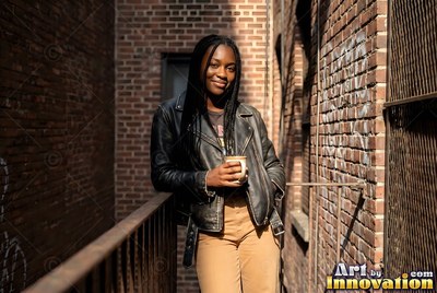 Holding a mug in the sun, a woman in a black leather jacket leans on a brick fire escape