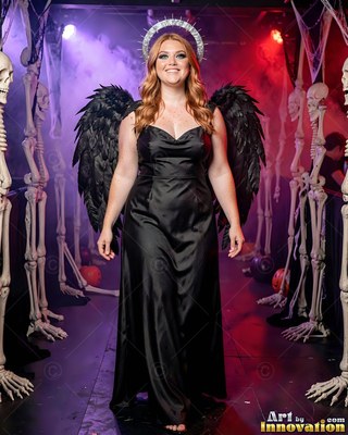 A grinning woman in a black gown and dark angel wings stands by skeletons