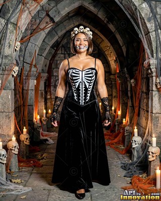 In a stone dungeon, a smiling woman models a black skeleton dress and a skull crown