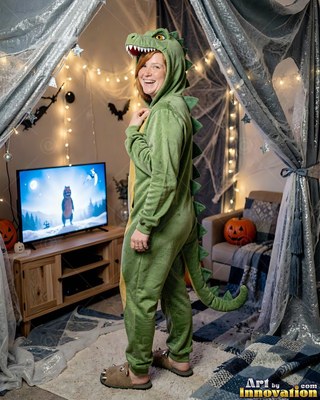 A woman in a green dinosaur onesie turns and smiles in a cozy living room