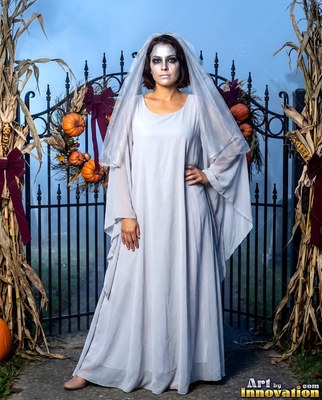 At an iron gate, a woman stands in a pale zombie bride gown and veil