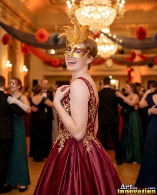 A woman smiling in a feathered gold mask and red gown in a ballroom