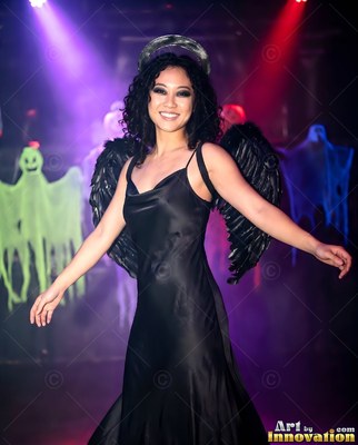 A woman smiling poses in a black angel dress with dark wings under party lights