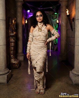 Posing in a stone tomb, a woman stands in a mummy costume with linen wraps and a gold scarab