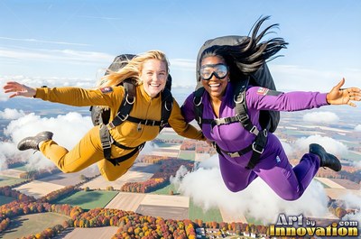 Two happy women skydive hand in hand over patchwork fields in bright daylight