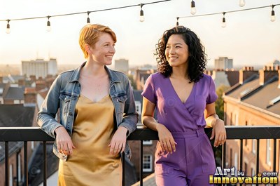 Leaning on a rooftop railing, two grinning women stand together in warm sunlight