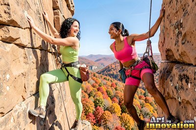Both women in bright green and pink gym clothes rock climb a cliff above autumn woods