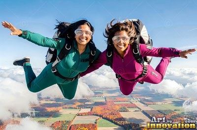 Two happy women in jumpsuits skydiving together above sunny clouds and fields