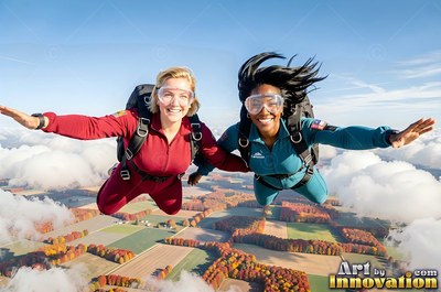 Wearing clear goggles, two woman friends skydive hand in hand high above patchy farmland in bright sun