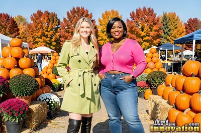 Dressed in a green coat and pink sweater, two adult women smile at a sunny pumpkin patch