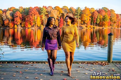 Walking side by side, two adult women laugh on a dock by a lake in fall