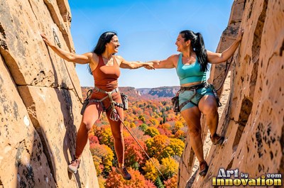 Two women rock climbing above trees with fall foliage and holding hands