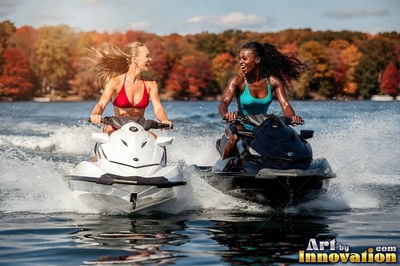 Both women sit atop jet skis across a lake under bright sunlight past orange trees