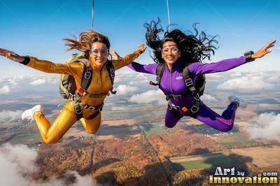 In the bright open sky, two woman friends skydive over fields in gold and purple jumpsuits