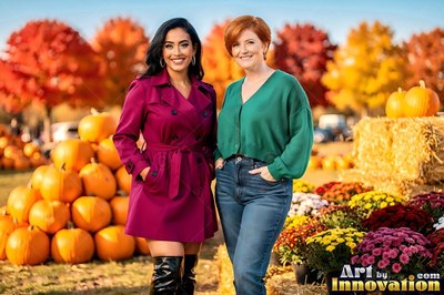 Two grinning women stand together outdoors by pumpkins in autumn sunlight