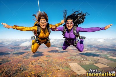 High in the blue sky, two smiling women skydive in yellow and purple jumpsuits