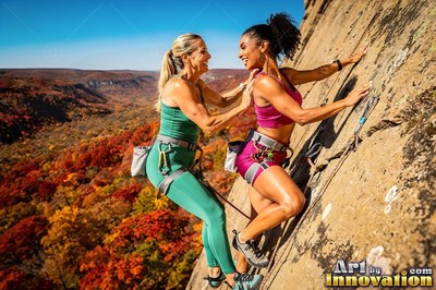 Climbing side by side, both women smile on a sunny cliff above fall trees