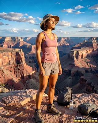 A woman in a brimmed sun hat and pink athletic tank top stands on a rocky canyon rim under the sun