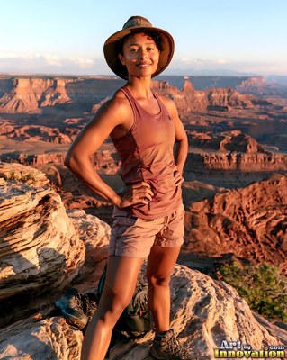 Standing on a rocky cliff, a smiling woman in a sun hat overlooks a red canyon edge