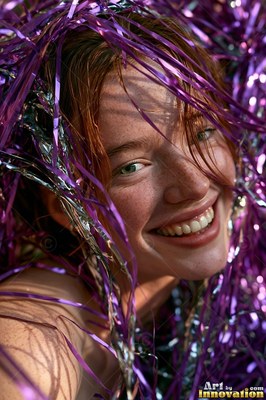 A smiling freckled woman peers out from under a pile of purple tinsel strands