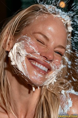 In bright light, a smiling blonde woman gets splattered with white cream