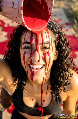 A cheerful woman in a black tank top gets red paint poured on her face outdoors