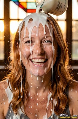A smiling redheaded woman has white milk poured over her head in warm sunlight