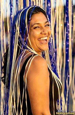 A laughing woman stands outdoors in sunlight with blue and white streamers draped over her dark hair