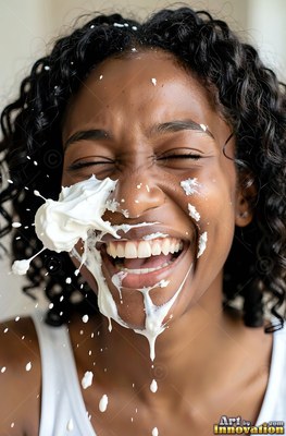 A smiling Black woman in a white tank top gets hit in the face with whipped cream