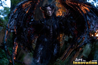 A woman bearing horns stands in a dark forest with black wings lit from behind by fire