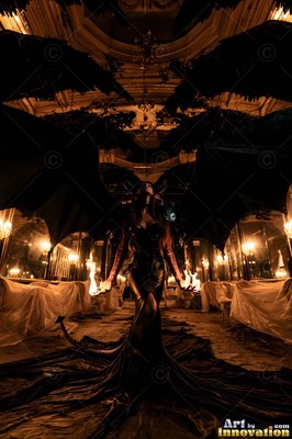 A winged woman in a dark dress holds fire in a candlelit ballroom