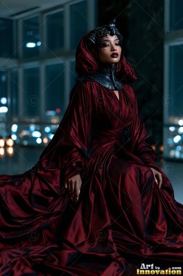 Before city windows at night, a woman sits draped in a dark red gown