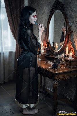 A light-skinned woman in a sheer black robe puts on makeup by candlelight at a vanity mirror